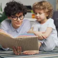 Seorang ibu sedang mendampingi dan mengajari anak membaca buku cerita bergambar dengan penuh keceriaan di rumah