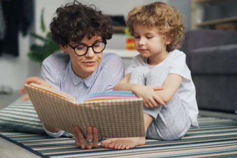 Seorang ibu sedang mendampingi dan mengajari anak membaca buku cerita bergambar dengan penuh keceriaan di rumah