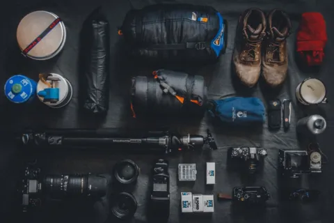 Perlengkapan traveler wajib bawa seperti paspor, kamera, kacamata, dan botol minum di atas meja kayu